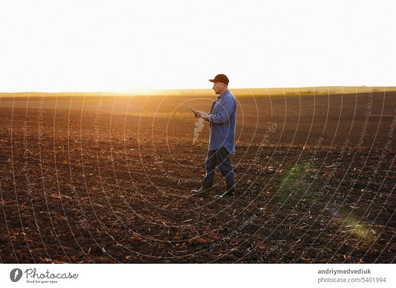 Intelligente Landtechnik und Landwirtschaft. Ein Landwirt benutzt ein digitales Tablet auf einem Feld mit gepflügtem Boden bei Sonnenuntergang. Überprüfung und Kontrolle der Bodenqualität, der Bereitschaft zur Aussaat von Pflanzen und zum Anbau von Gemüse