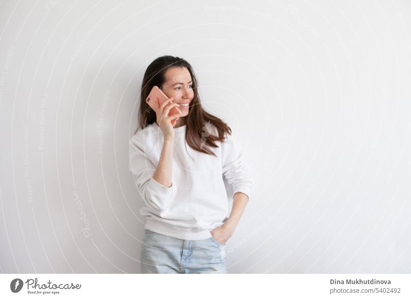 Junge Frau hält ein Mobiltelefon, lächelnd, mit sozialen Medien. Millennial Person in weißem Sweatshirt auf weißem Hintergrund. Menschen nutzen Technologie. Anruf bei der Arbeit.