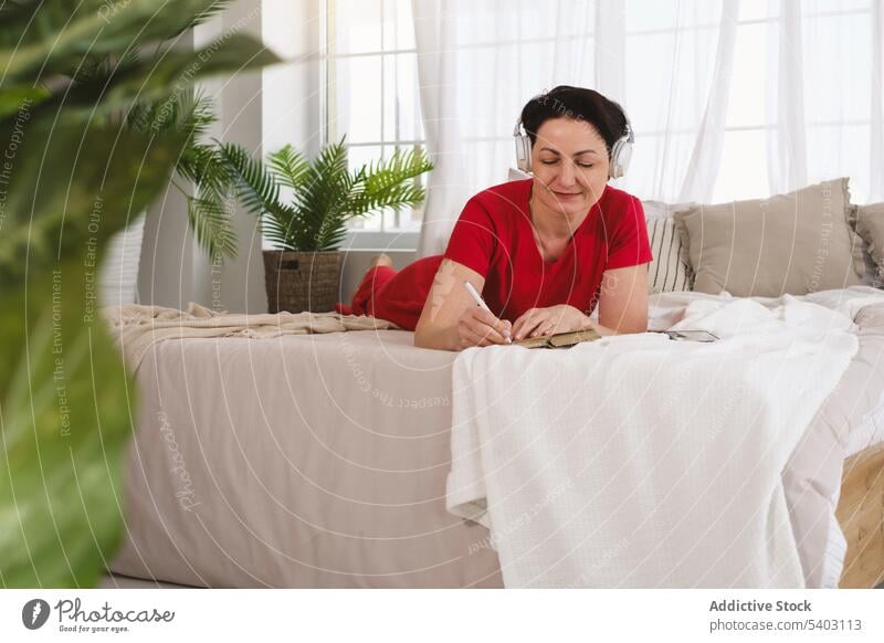 Frau hört Musik und schreibt Notizen auf dem Bett Fokus Konzentration schreiben Notebook zur Kenntnis nehmen zuhören Kopfhörer Schlafzimmer Lügen Laptop Gesang