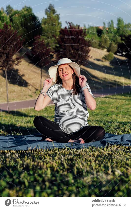 Schwangere Frau auf Decke sitzend schwanger erwarten Bauch mütterlich Lotus-Pose grün Rasen Augen geschlossen Glück Hut Schwangerschaft Kinderkriegen pränatal