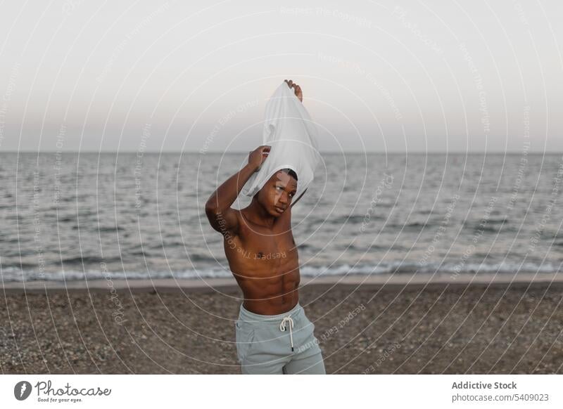 Schwarzer Sportler beim Ausziehen des T-Shirts am Strand stehend Mann muskulös Macho nackter Torso passen stark ohne Hemd Figur brutal männlich sportlich Körper