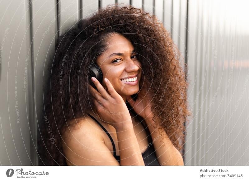 Charmante ethnische Frau mit Kopfhörer auf der Straße krause Haare Afro-Look Frisur charmant Lächeln natürlich Schönheit zuhören schwarz Afroamerikaner