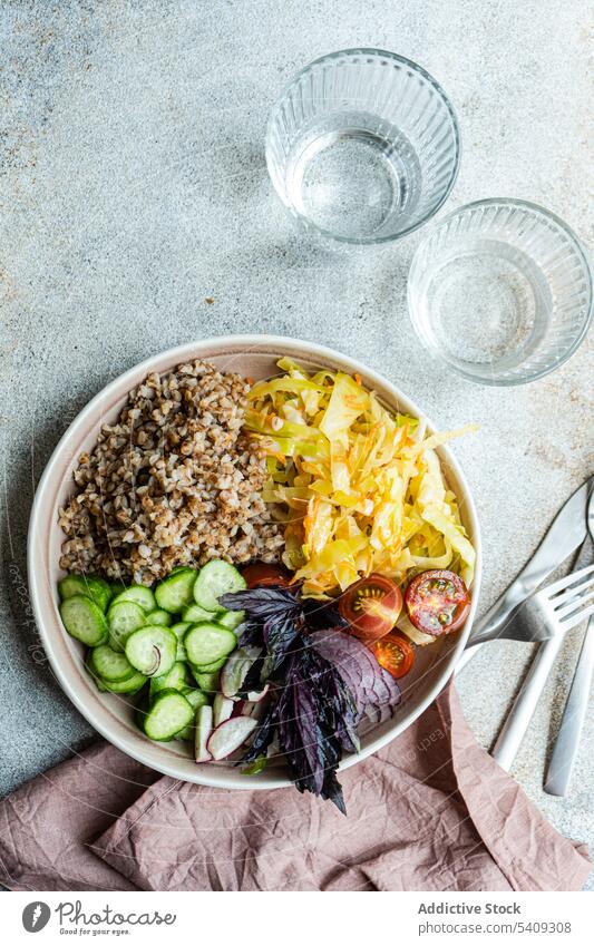 Teller mit leckerem Veggie-Gericht, Besteck und Gläser auf dem Tisch Vegetarier Buchweizen Gemüse Zwiebel Salatgurke Tomate Basilikum Schalen & Schüsseln