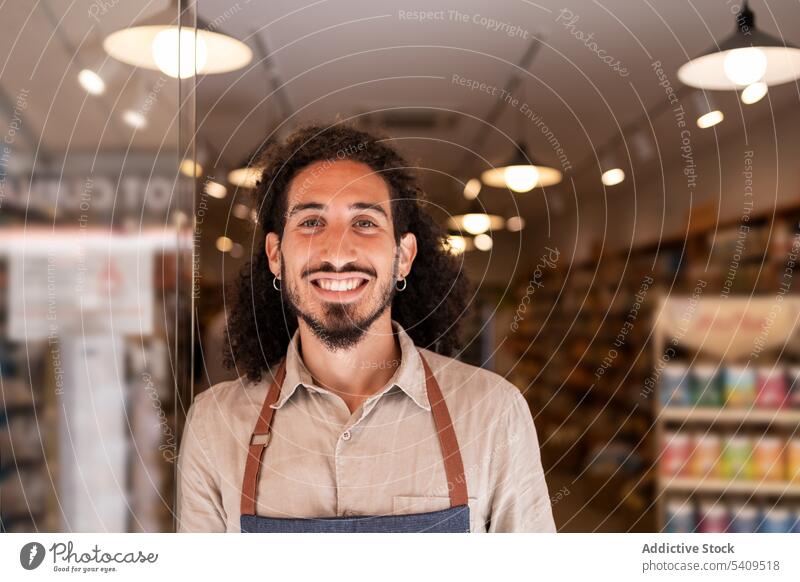 Fröhlicher Mann, der vor einem beleuchteten Supermarkt mit Waren steht Lächeln selbstbewusst Porträt Vollbart positiv heiter Glück Schürze Arbeit freundlich