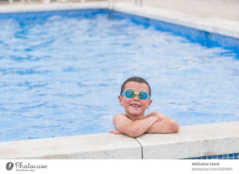 Glückliches Kind mit Schwimmbrille im Schwimmbad stehend bei Tageslicht Lächeln Schutzbrille Pool nasses Haar Wasser genießen froh die Hände gekreuzt Beckenrand