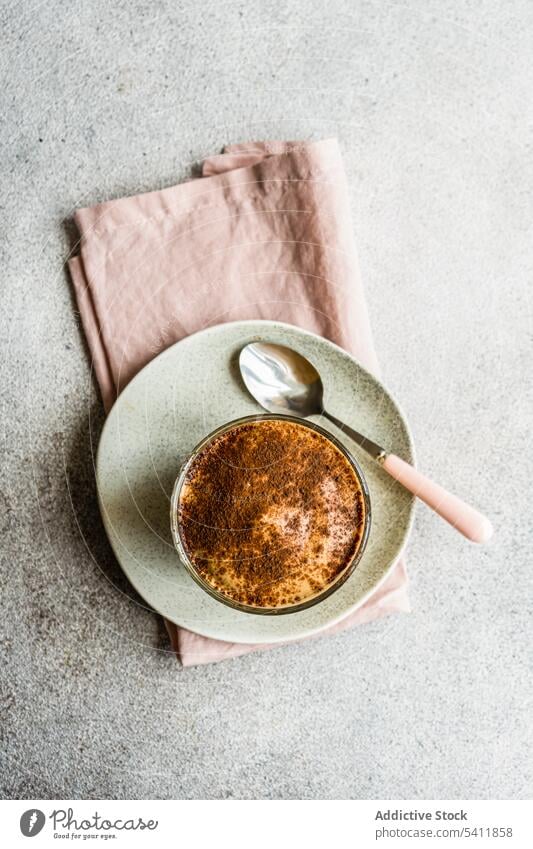 Glasbecher mit Kaffeesahne-Dessert mit Schokoladenpulver und Eis Tasse Becher Pulver Löffel Teller Serviette Tisch Oberfläche grau verschwommener Hintergrund