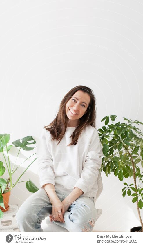 Millennial Frau trägt weißes Sweatshirt und Jeans, lächelnd, sitzt auf einem Stuhl zu Hause mit grünen Pflanzen. Hipster Lebensstil der Millennials. Lässiges Porträt.