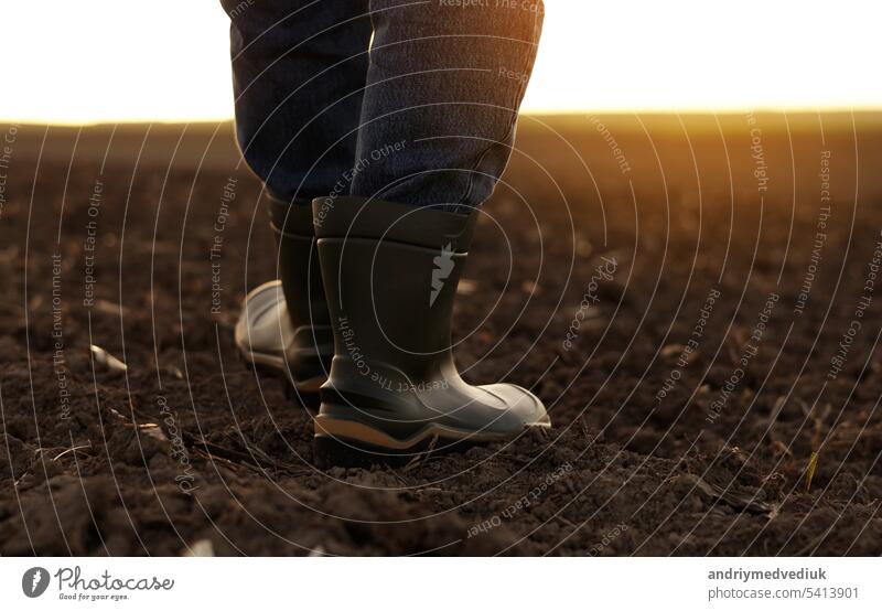 Landwirtschaft. Cropped Schuss von hinten Blick Geschäftsmann Landwirt in Gummistiefeln geht entlang gepflügten Feld. Agronom überprüft und analysiert fruchtbaren Boden bei Sonnenaufgang. Agribusiness.