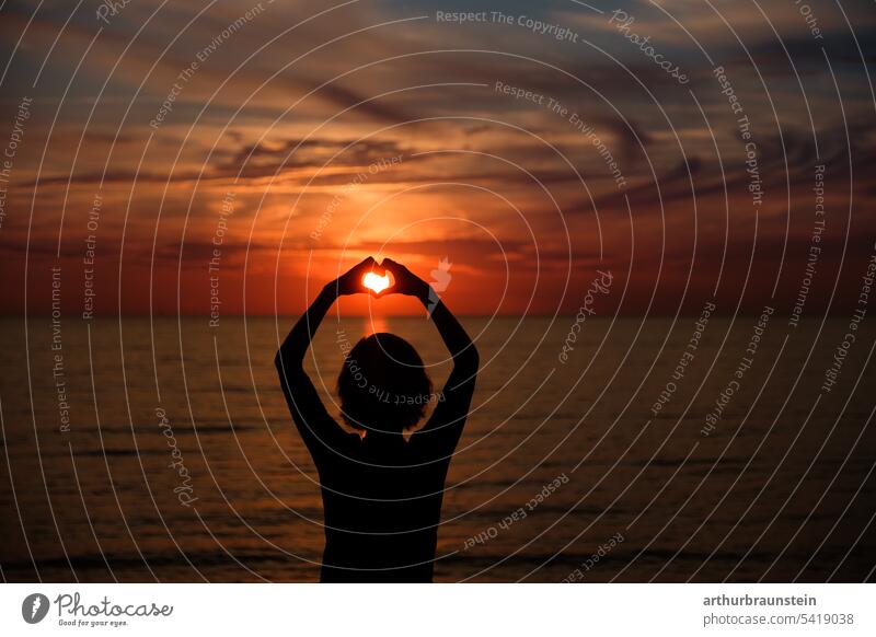 Junge Frau auf Felsen formt Herz mit den Händen bei Sonnenuntergang am Meer Sonnenlicht sonnig Sommer Sommerurlaub sommerlich Sommerzeit Sommergefühl Meerwasser