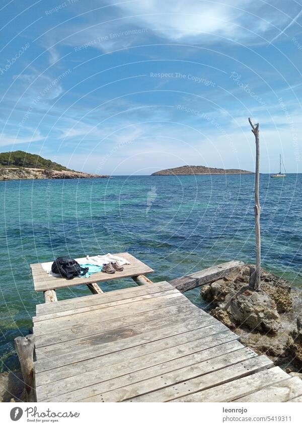 Ein Holzsteg mit Blick auf das Meer und die Wellen auf der Mittelmeerinsel Ibiza. Im Hintergrund sieht man eine kleine Insel und ein Segelboot.