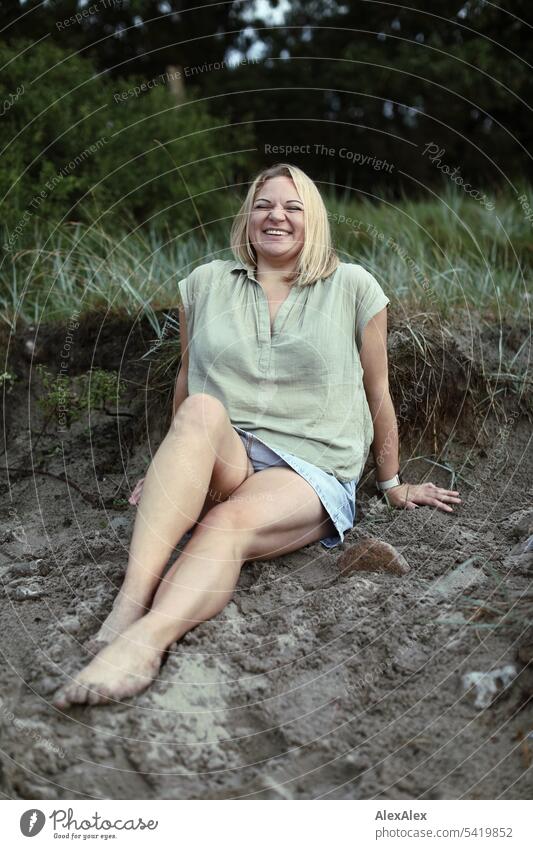 Portrait einer schönen, blonden Frau die im Sand am Strand sitzt und sich freut langhaarig blonde Haare Freude Lächeln Dünengras Schönheit entspannt relaxed