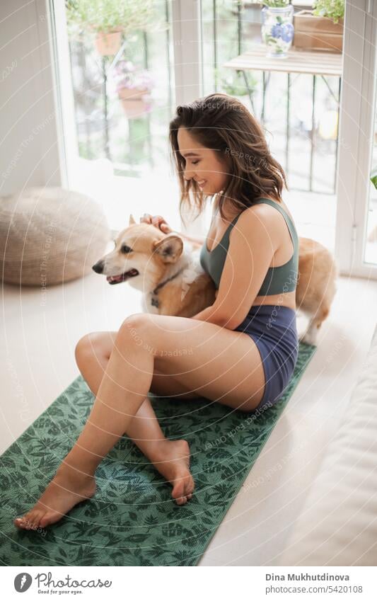 Junge Frau mit Corgi Pembroke Hund macht Yoga auf grüner Matte zu Hause. Praktiziert Sport, Wellness und Gesundheit. Indoor Yoga Online-Kurs. Achtsamkeit und Meditation, aktiver Lebensstil.