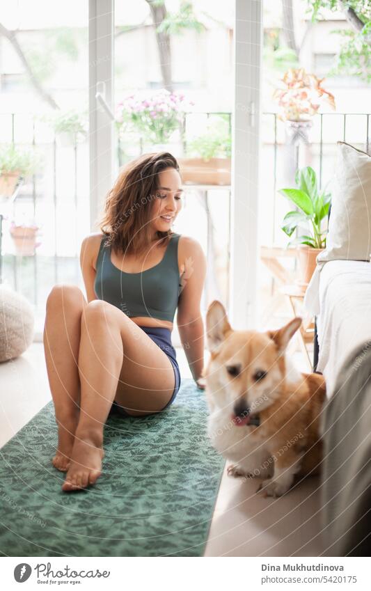 Junge Frau mit Corgi Pembroke Hund macht Yoga auf grüner Matte zu Hause. Praktiziert Sport, Wellness und Gesundheit. Indoor Yoga Online-Kurs. Achtsamkeit und Meditation, aktiver Lebensstil.