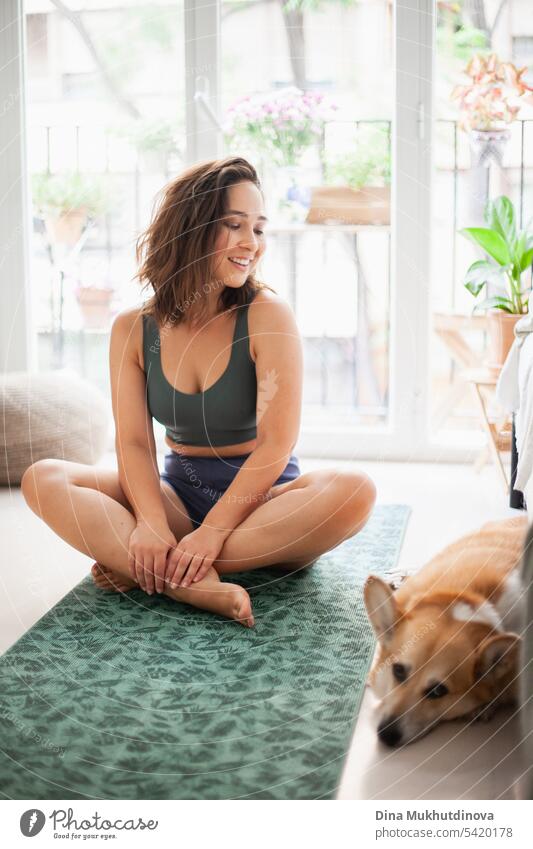 Junge Frau mit Corgi Pembroke Hund macht Yoga auf grüner Matte zu Hause. Praktiziert Sport, Wellness und Gesundheit. Indoor Yoga Online-Kurs. Achtsamkeit und Meditation, aktiver Lebensstil.