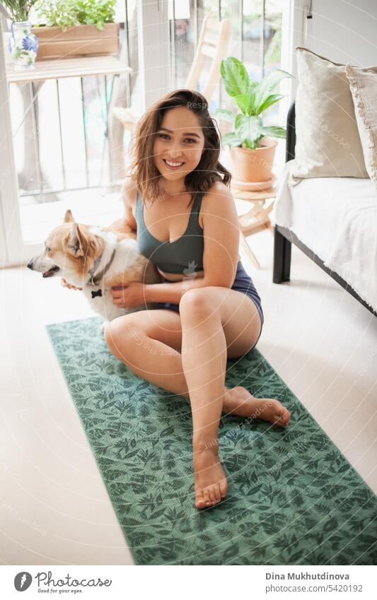 Junge Frau mit Corgi Pembroke Hund macht Yoga auf grüner Matte zu Hause. Praktiziert Sport, Wellness und Gesundheit. Indoor Yoga Online-Kurs. Achtsamkeit und Meditation, aktiver Lebensstil.