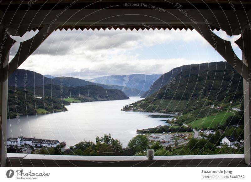 Morgenkaffee mit schönem Blick auf einen Fjord und Berge in Norwegen von der Veranda aus. Im Hintergrund sieht man einen Fjord mit einem kleinen Dorf. Der Himmel ist bewölkt.