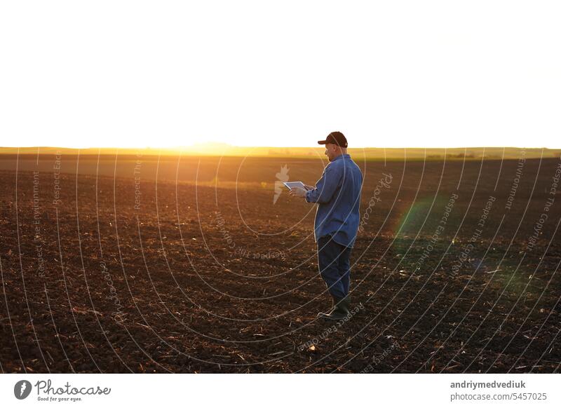 Intelligente Landtechnik und Landwirtschaft. Ein Landwirt benutzt ein digitales Tablet auf einem Feld mit gepflügtem Boden bei Sonnenuntergang. Überprüfung und Kontrolle der Bodenqualität, der Bereitschaft zur Aussaat von Pflanzen und zum Anbau von Gemüse