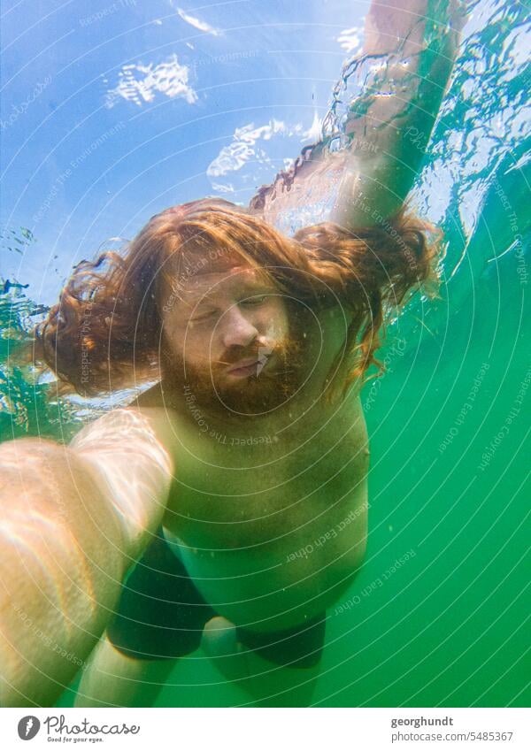 Mann mit rotem Haar und rotem Bart taucht in einem Süßwassersee. Die Augen offen, aber leicht zugekniffen. Er hält die Kamera. See unterwasser tauchen schwimmen