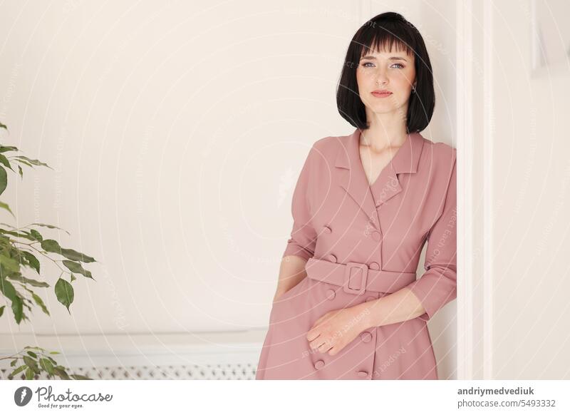 Porträt der attraktiven lächelnden Brünette mit kurzen Haaren Frau allein auf weißem stilvollen Wand Hintergrund mit grünen Zimmerpflanze bleiben. Weiblich in weichen rosa Kleid ist in der Kamera suchen