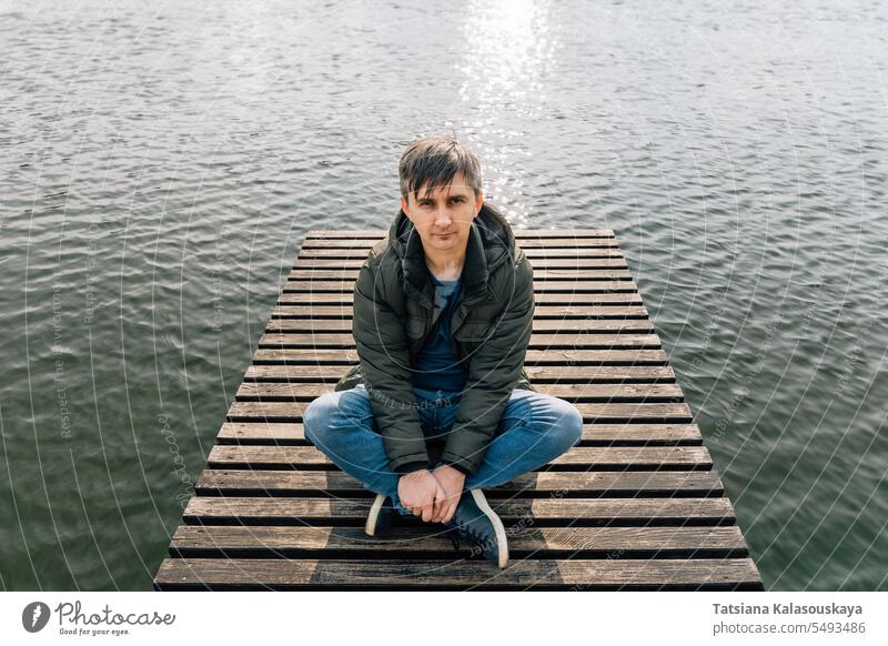 Ein Mann sitzt entspannt auf einem Holzsteg am See, im Schneidersitz, in Jeans und Jacke im Herbst Jeanshose Trägerkleid sitzen entspannend hölzern