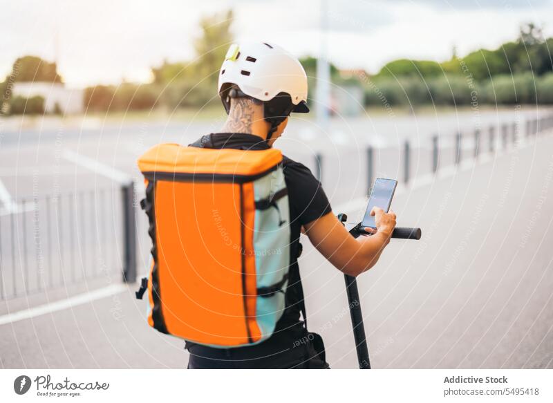 Anonymer männlicher Kurier, der ein Handy auf einem Kick-Scooter benutzt Auslieferer Smartphone benutzend elektrisch Kickroller Schutzhelm Rucksack navigieren