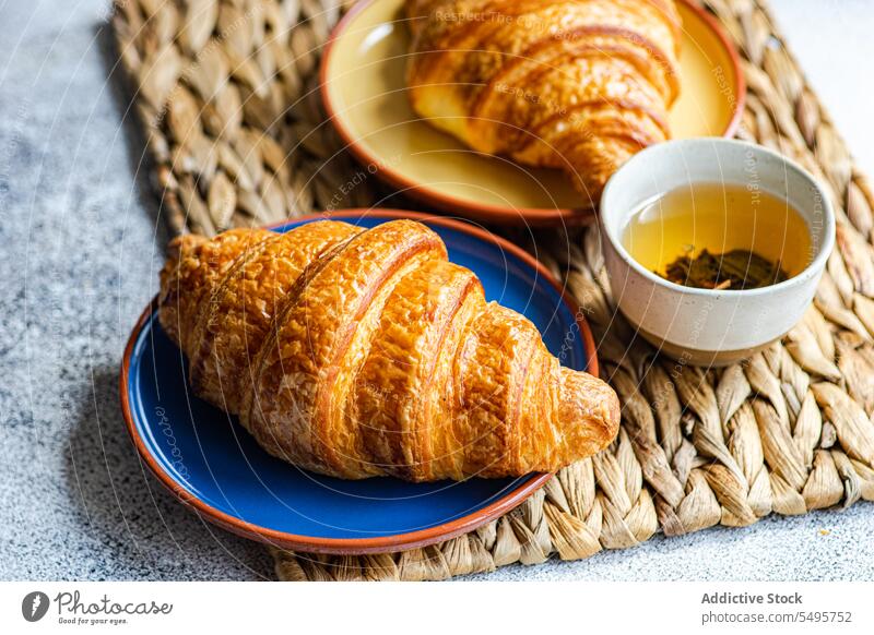 Frisch gebackene Croissants auf Keramiktellern frisch Teller farbenfroh braun Serviette Tasse grün Tee Tisch Oberfläche grauer Hintergrund hoher Winkel von oben