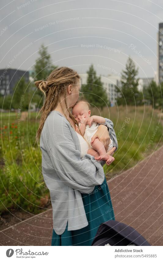 Frau trägt und küsst Baby im Park gegen den Himmel Mutter tragen Küssen Kopf Liebe klein Umarmung bezaubernd Familie Lifestyle niedlich Rastalocken Säugling