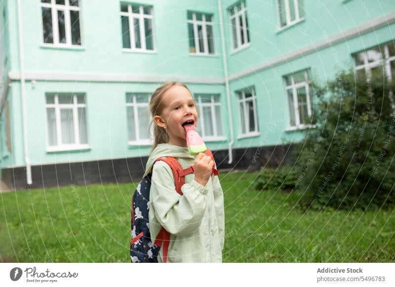 Glückliches Kind mit Rucksack im grünen Park stehend und ein Eis im Tageslicht genießend Speiseeis Kindheit Gras essen niedlich bezaubernd Mädchen Feld