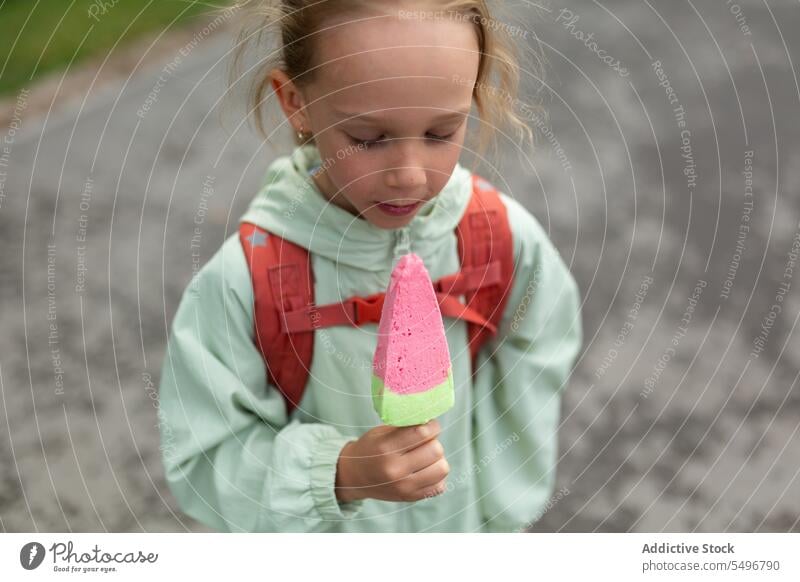 Mädchen mit Rucksack steht in der Nähe grasbewachsenen Feld und genießt Eis Kind Speiseeis Park Weg niedlich Kindheit grün bezaubernd Sommer süß sorgenfrei