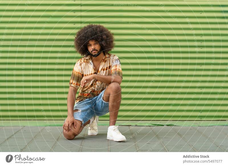 Stilvoller ethnischer Mann mit Afro-Haar vor gestreifter Wand trendy selbstbewusst Afro-Look cool krause Haare unabhängig Individualität hocken Outfit Vorschein