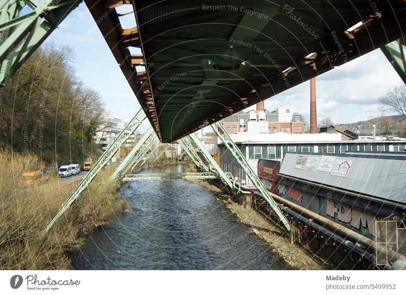 Stahlträger der Gleise der Wuppertaler Schwebebahn über der Wupper mit alter Fabrik bei Sonnenschein im Stadtzentrum von Wuppertal im Bergischen Land in Nordrhein-Westfalen, Deutschland