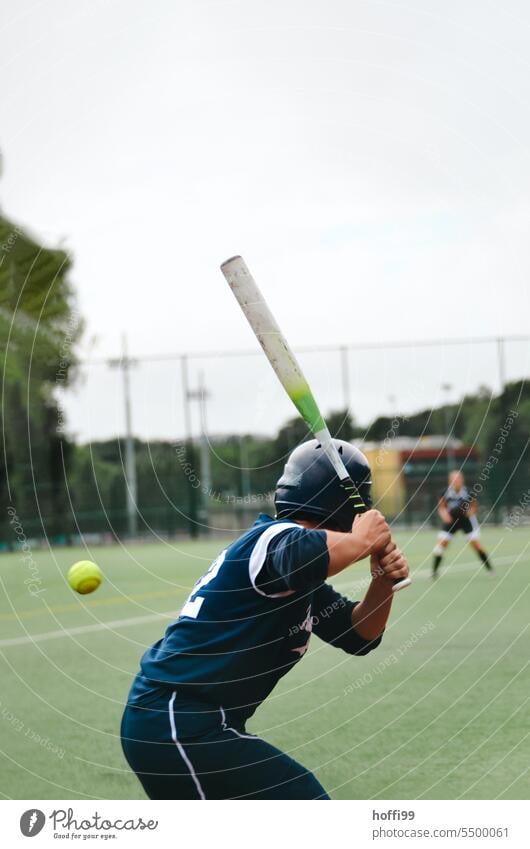 Softballspielerin / Baseballspieler mit dem Rücken zur Kamera hat einen heranfliegenden Ball fest fixiert und holt aus zum Schlag um ihn zu treffen Rasen