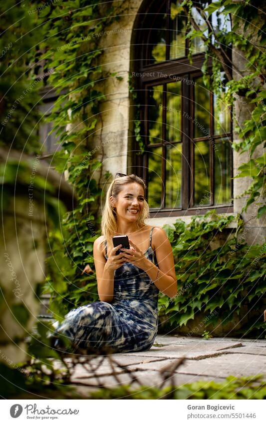 Hübsche junge Frau, die ein Handy benutzt, neben einem alten Haus mit Efeu Sonnenbrille Sitzen niedlich Kaukasier Erwachsener attraktiv schön Anruf Funktelefon