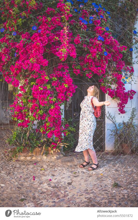 blonde Frau mittleren Alters mit Kleid steht unter einer riesigen purpur blühenden Pflanze an einem Haus in Spanien Sommer Andalusien Granada Bougainvillea lila