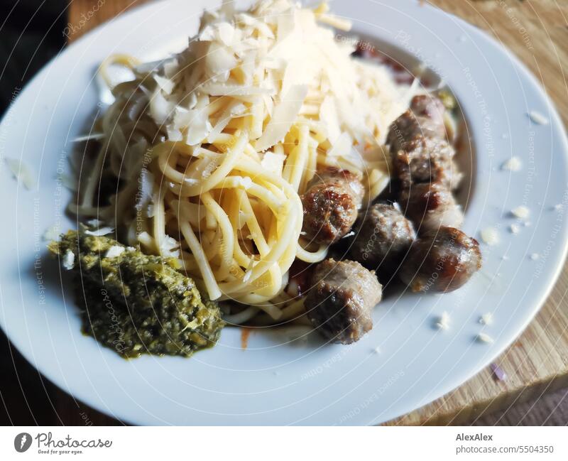 Spaghetti mit Bratwürstchen, Parmesanflocken und grünem Pesto auf einem weißen Teller, der auf einem hölzernen Schneidebrett steht Pasta Nudeln Teigwaren