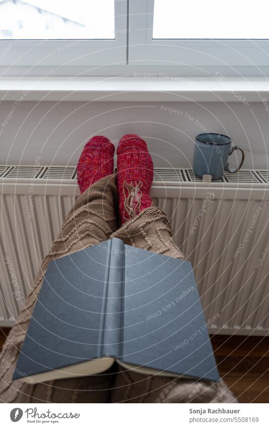 Eine Frau mit Wollsocken und einer Wolldecke wärmt sich an einem Heizkörper Beine Wärme Buch gemütlich Hygge heizen Winter kalt Heizung Heizkosten Temperatur