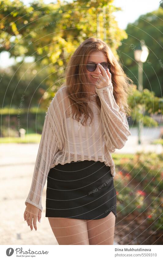 candid Porträt der jungen schönen Frau lachend in Sonnenbrille im Gegenlicht mit Sonnenlicht im Sommer. Hübsches rothaariges Mädchen mit roten Haaren. Park