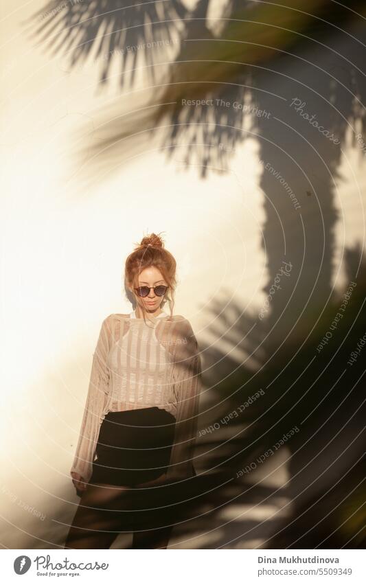 junge schöne rothaarige Frau in Freizeitkleidung mit Sonnenbrille und Haarknoten auf beigem Wandhintergrund mit Palmenschatten. Candid Outdoor-Porträt im Park.