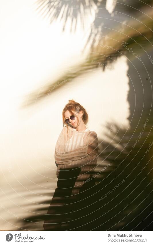 junge schöne rothaarige Frau in Freizeitkleidung mit Sonnenbrille und Haarknoten auf beigem Wandhintergrund mit Palmenschatten. Candid Outdoor-Porträt im Park.