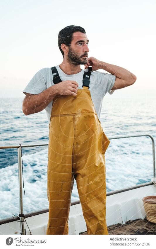 Selbstbewusster junger ethnischer Fischer, der die Hosenträger seines Lätzchens auf dem Segelboot festzieht Mann Boot reisen befestigen wasserdicht Klammer