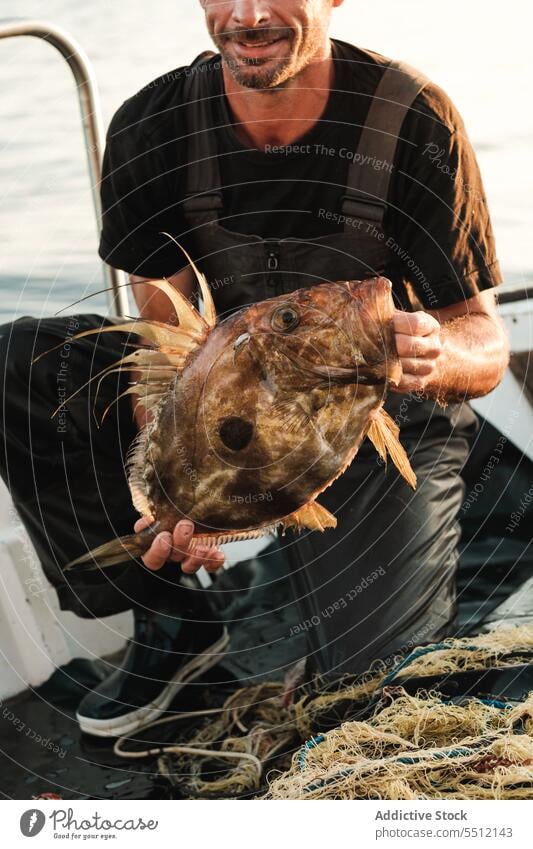 Crop Mann sitzt auf Bootsdeck und hält Fisch mit großen Mund im Tageslicht Schwimmer Schiffsdeck marin Wasser Verkehr Segel Sommer Natur männlich Meer Fischen