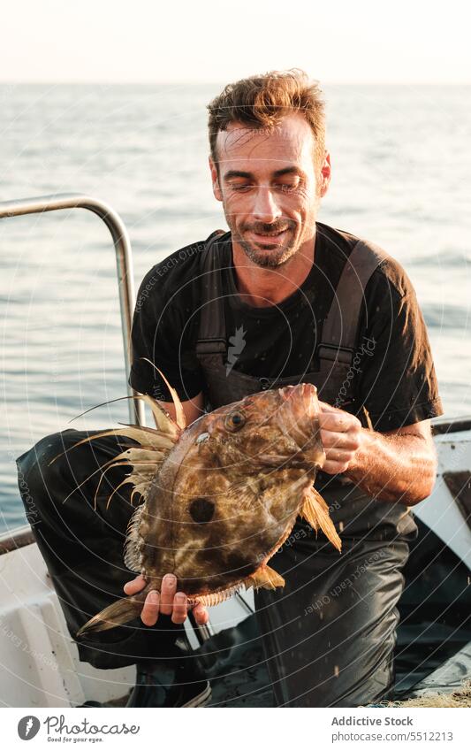 Glücklicher Mann sitzt auf dem Bootsdeck und hält einen Fisch mit großem Maul im Tageslicht Schwimmer Schiffsdeck marin Lächeln Wasser Verkehr Freude Segel