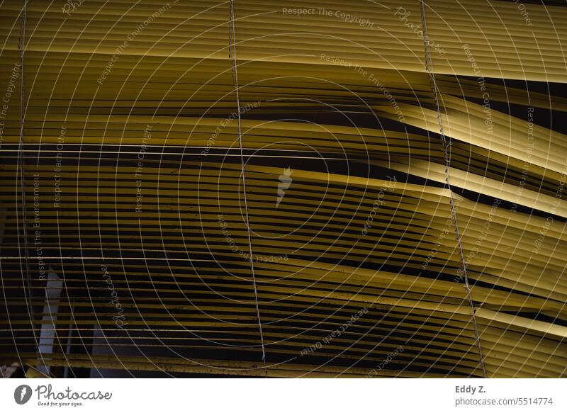 Eingedrückte Lamellen einer gelben Jalousie vor einem dunklen Raum, der neugierige Blicke von draußen herausfordert. Lamellenjalousie Fenster Sichtschutz