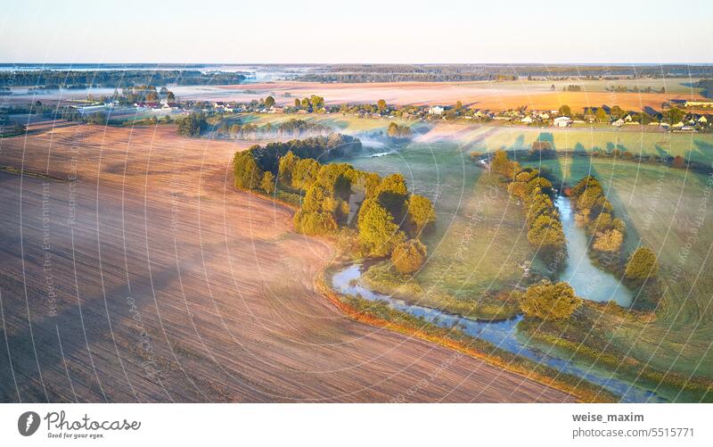 Warmer sonniger Septembermorgen. Herbstlich schöne Nebellandschaft. Kleiner Fluss Kurven in Bäumen. Helles sonniges nebliges Flussufer. Scenic Ansicht der ländlichen Sonnenaufgang. Dorf in der Landwirtschaft Felder