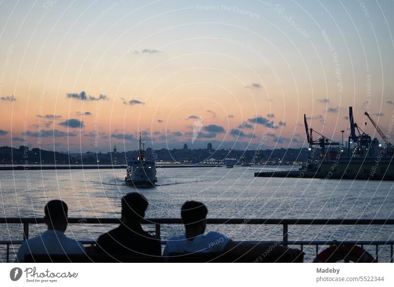 Touristen an Deck einer Fähre über den Bosporus am Containerhafen Haydarpasa mit romantischem Sonnenuntergang im Stadtteil Kadiköy in Istanbul in der Türkei