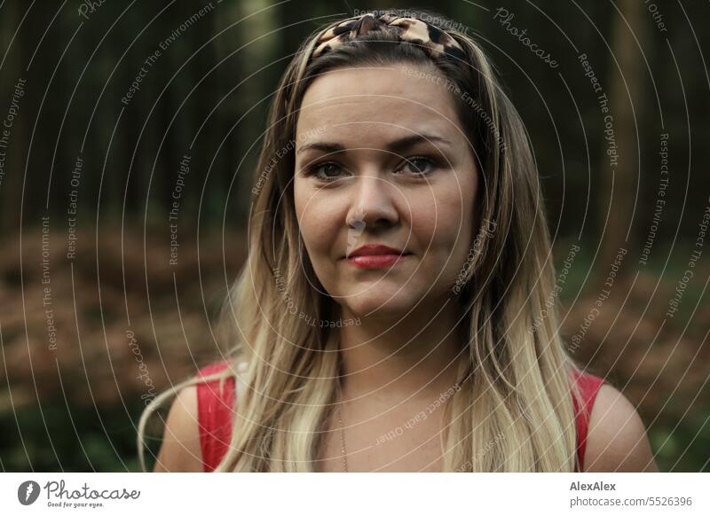 Portrait einer schönen, blonden, langhaarigen Frau, die in einem lichten Wald steht und in die Kamera schaut blonde Haare lange Haare rotes Kleid Pflanze grün