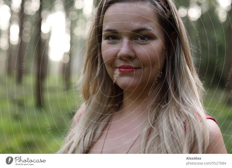 Portrait einer schönen, blonden, langhaarigen Frau, die in einem lichten Wald steht und in die Kamera schaut blonde Haare lange Haare rotes Kleid Pflanze grün