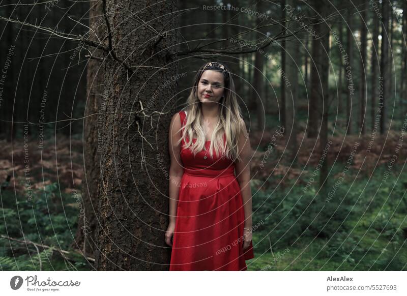 Portrait einer schönen, blonden, langhaarigen Frau, die in einem lichten Wald an einem Baum gelehnt steht und in die Kamera schaut blonde Haare lange Haare