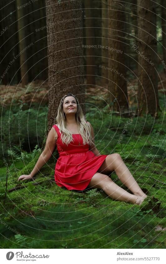 Portrait einer schönen, blonden, langhaarigen Frau, die in einem lichten Wald an einem Baum gelehnt sitzt und nach oben schaut blonde Haare lange Haare