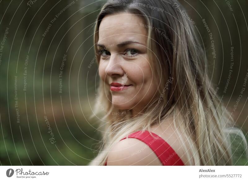 Portrait einer schönen, blonden, langhaarigen Frau, die in einem lichten Wald steht und nach hinten in die Kamera schaut blonde Haare lange Haare rotes Kleid